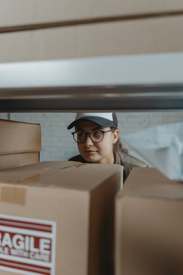 A woman in glasses and a cap peering over stacked cardboard boxes on a shelf.